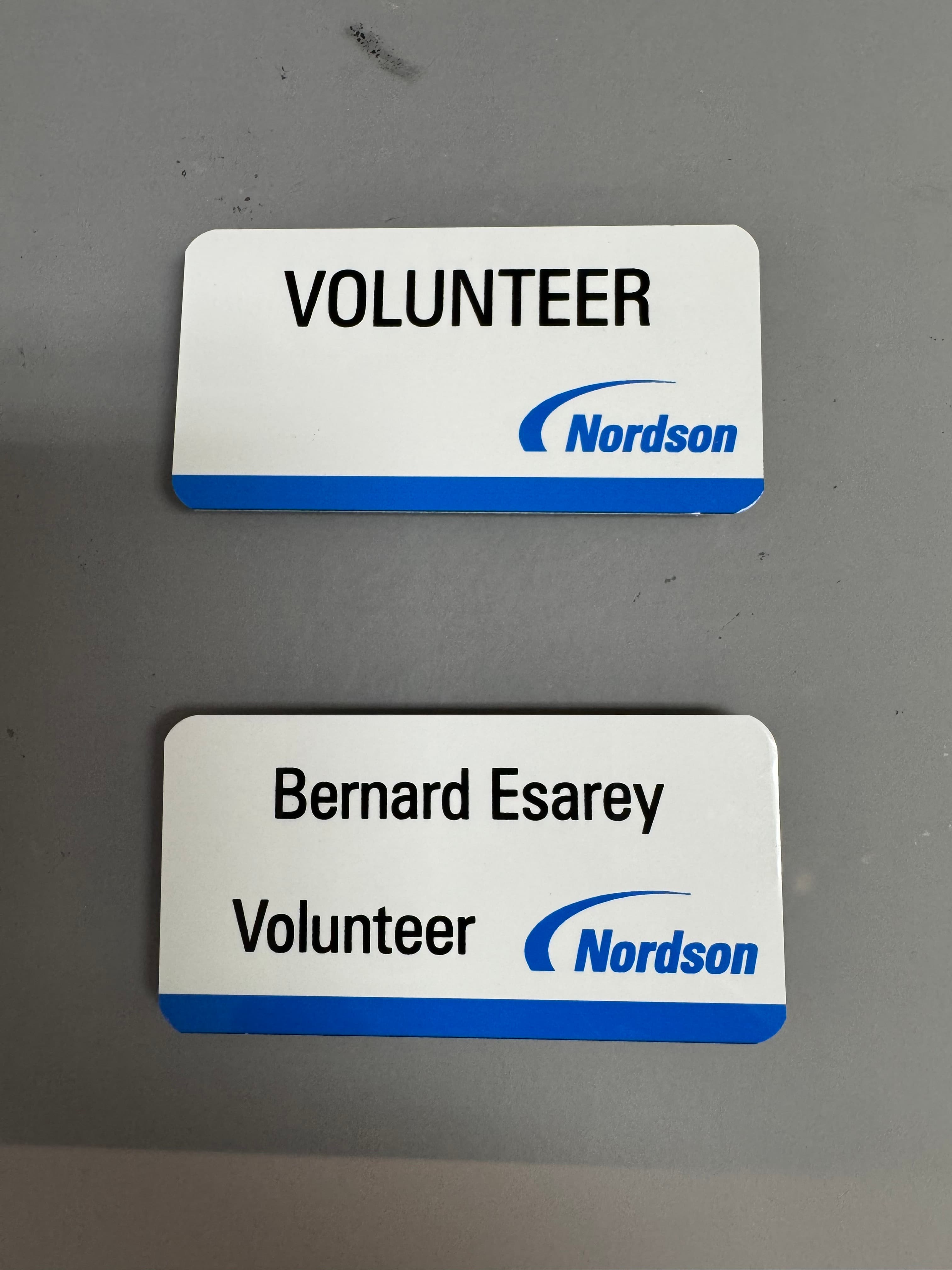Two white Nordson volunteer name tags, one for Bernard Esarey, resting on a grey surface.