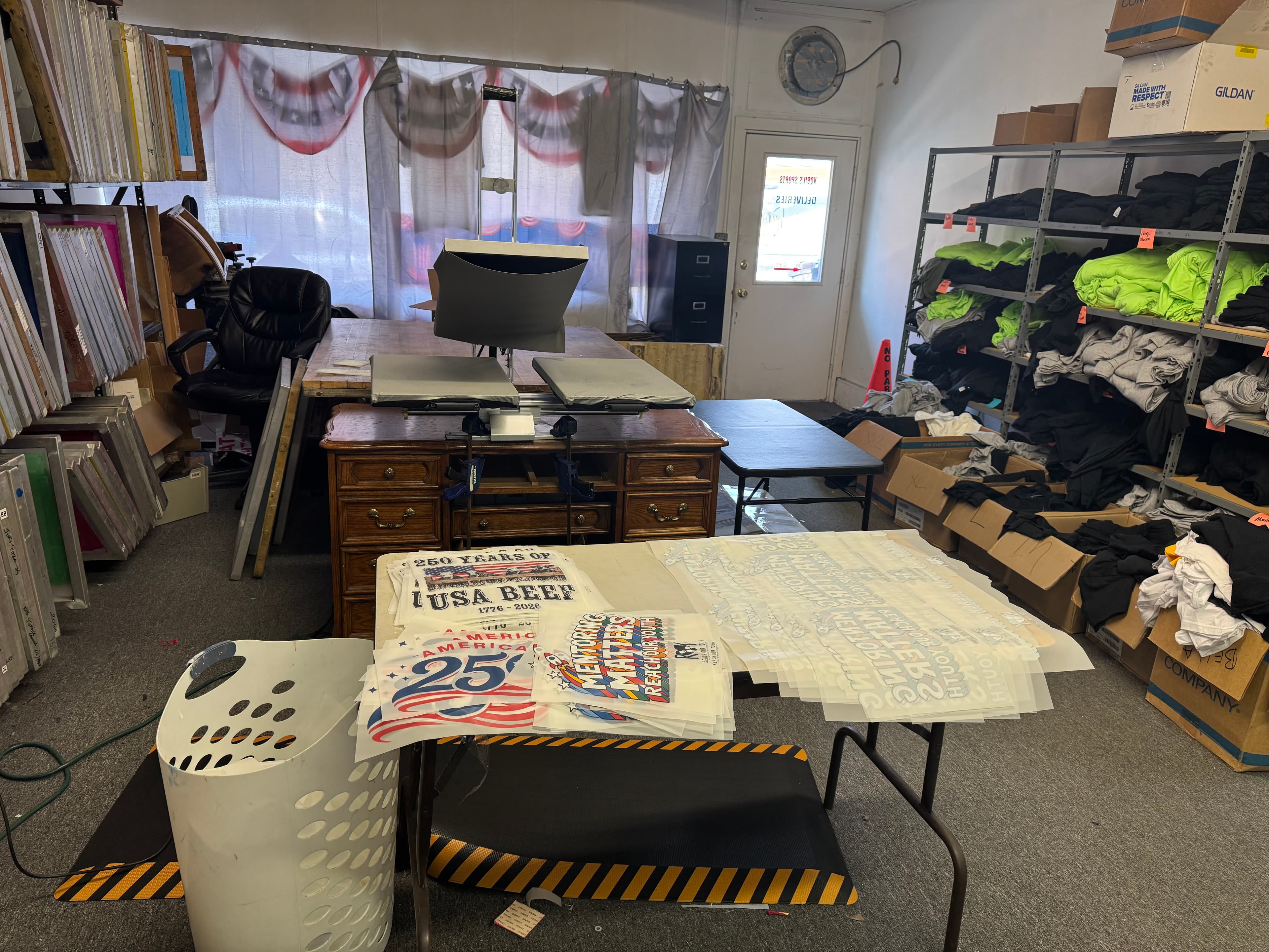Screen printing workshop with a heat press, printed transfers, and shelves of folded t-shirts.