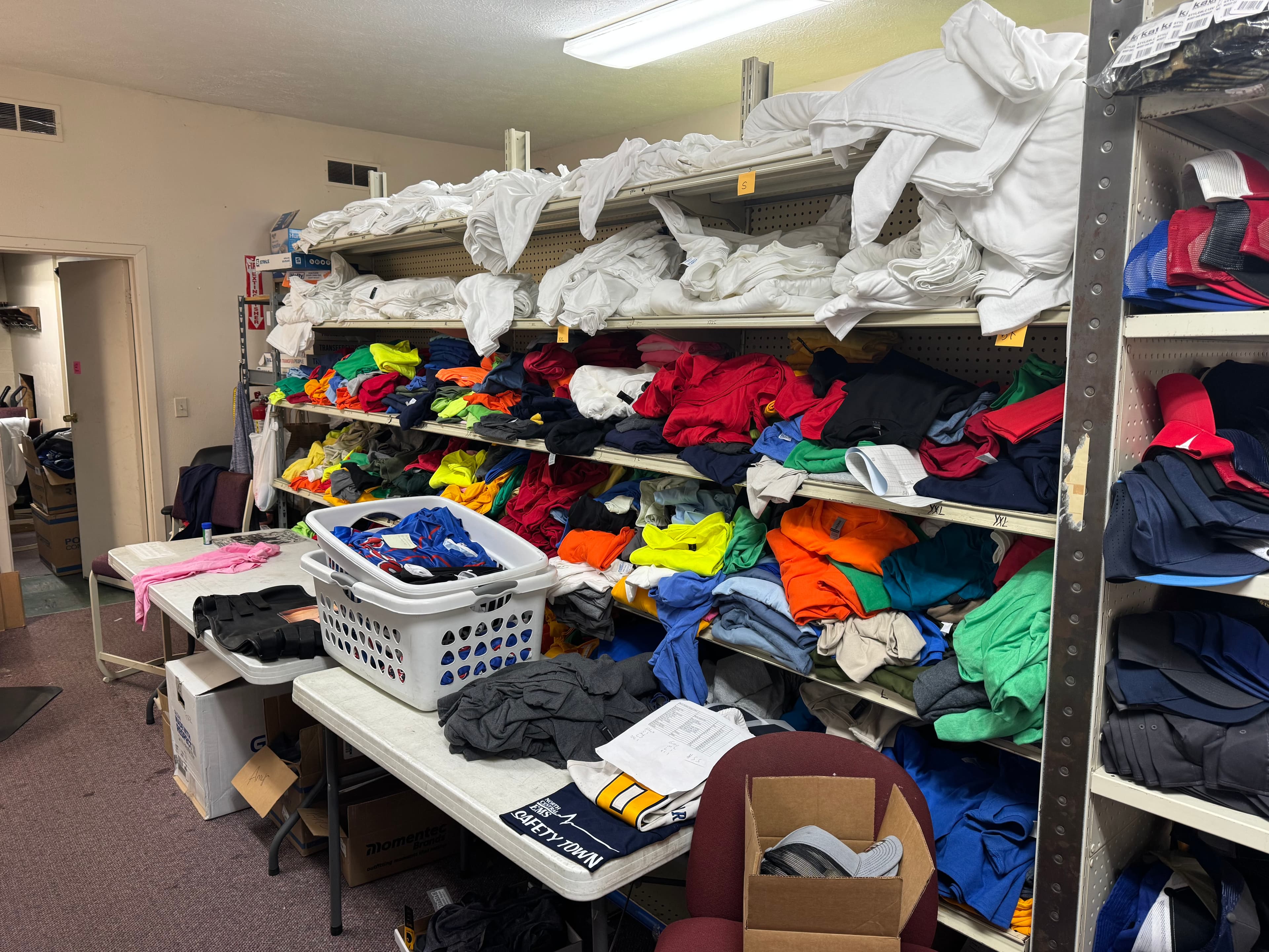 Metal shelves overflowing with colorful folded shirts and white laundry baskets in a storage room.