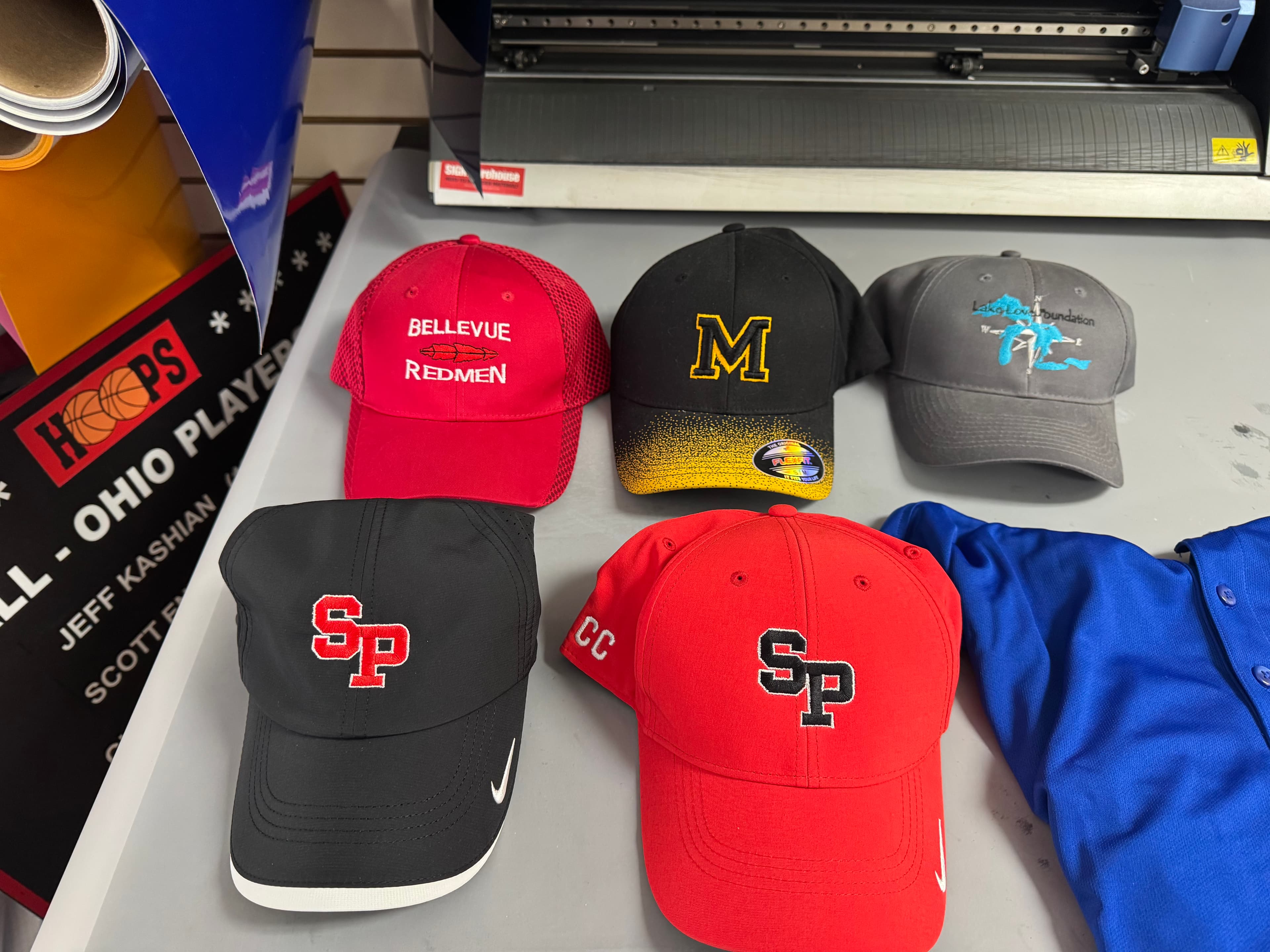 Five embroidered baseball caps with various logos displayed on a gray work surface.