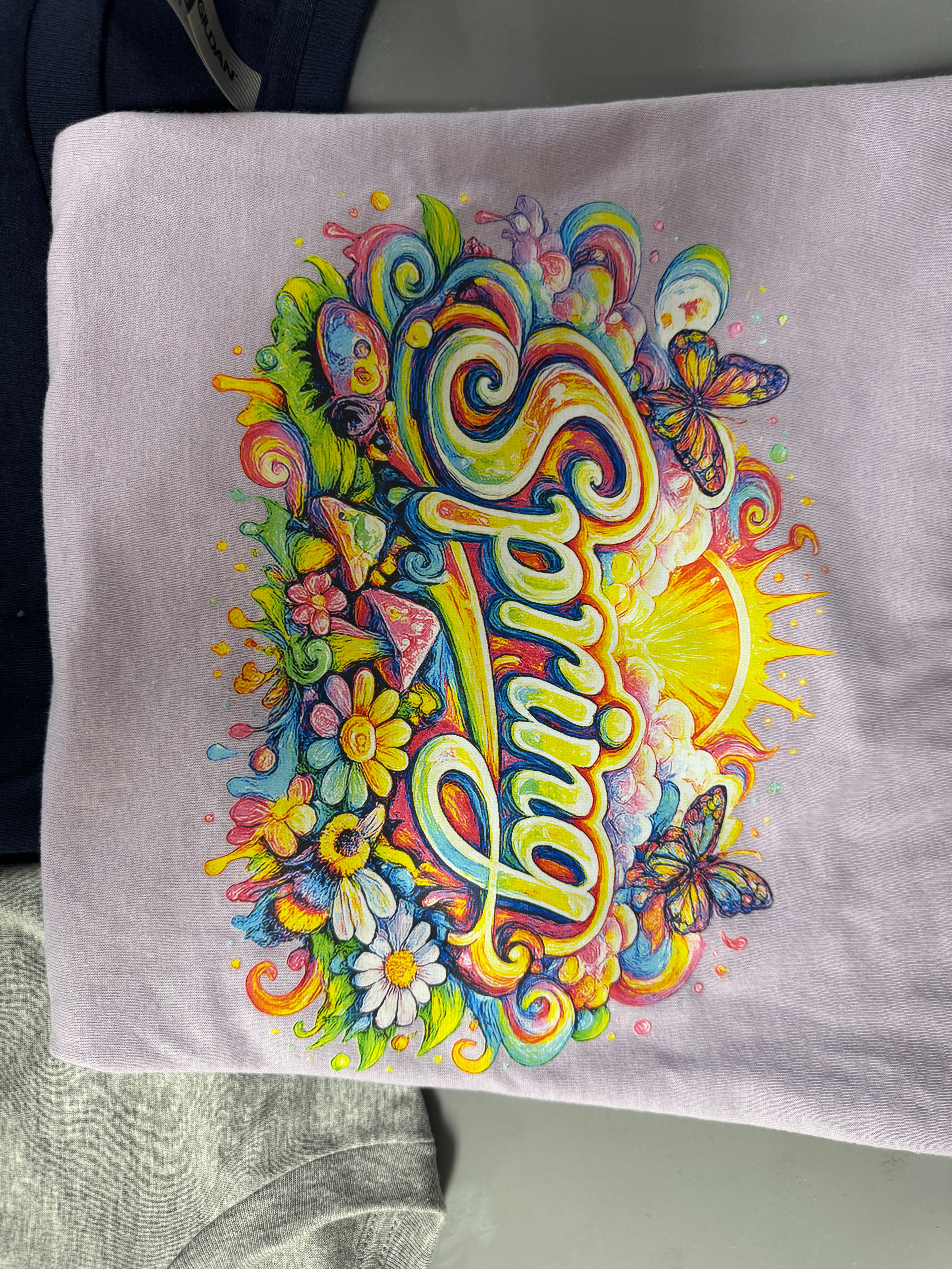 Vibrant psychedelic Spring graphic on a purple shirt with flowers, butterflies, and a bright sun.