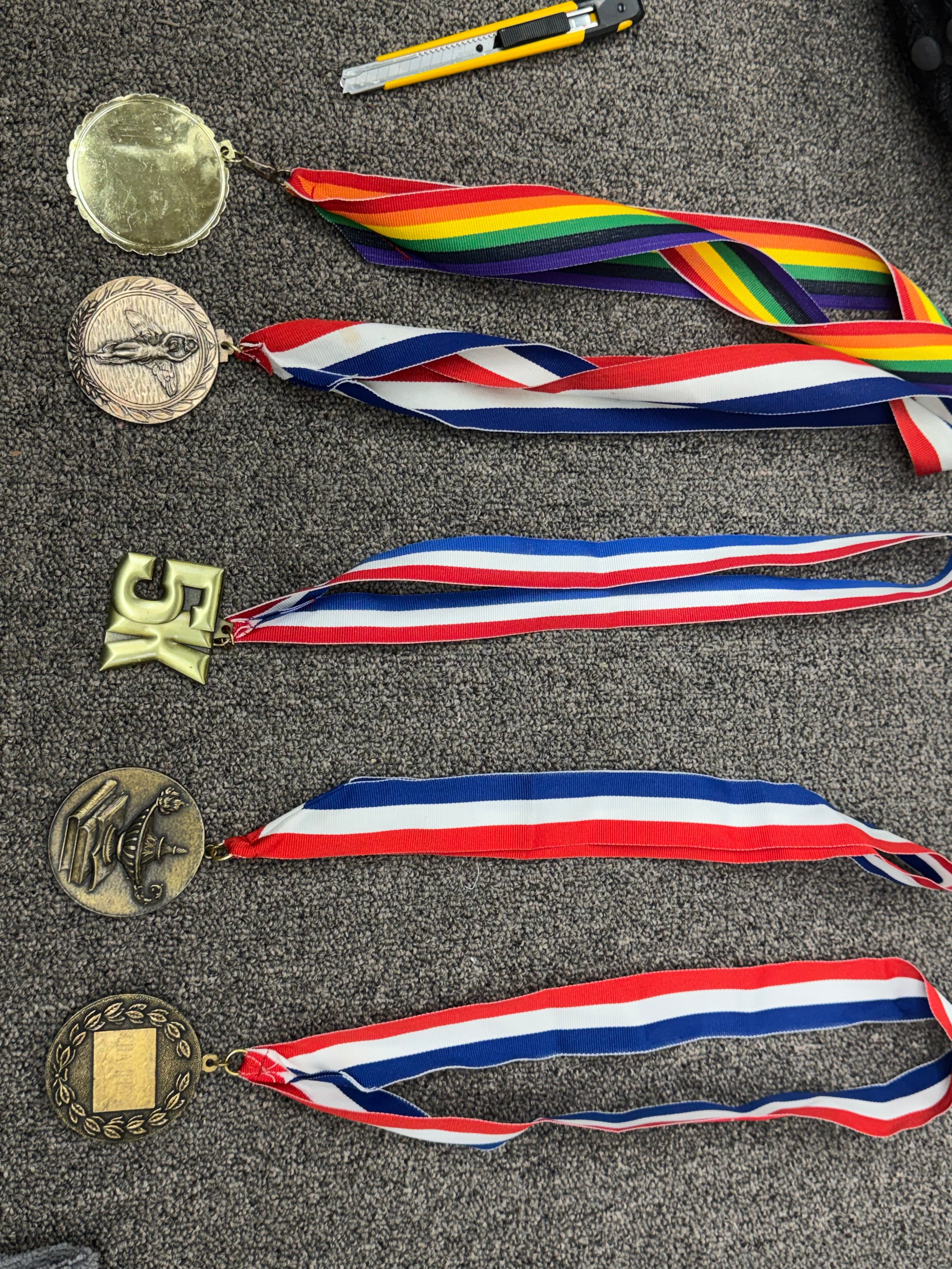 Five medals with rainbow and striped ribbons, including a 5K award, on grey carpet.