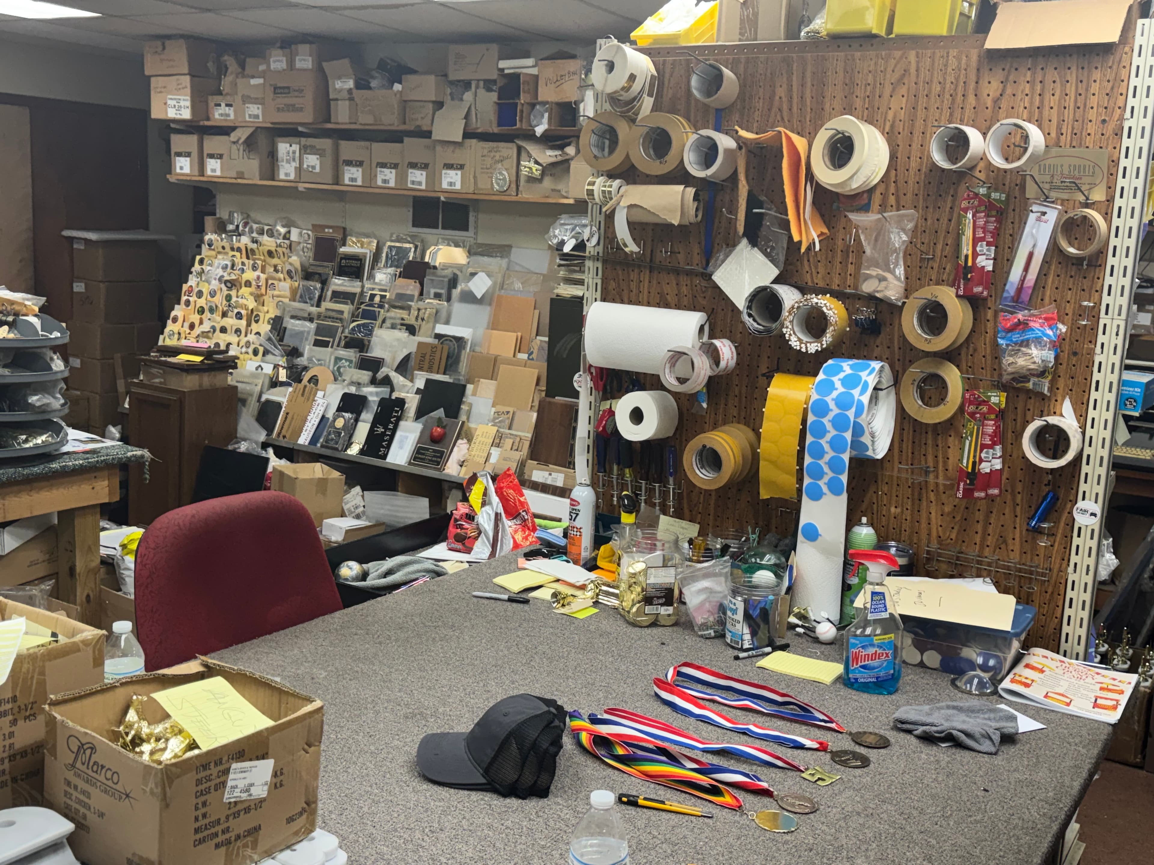 Cluttered workshop table with medals and a cap, backed by a pegboard and boxes.