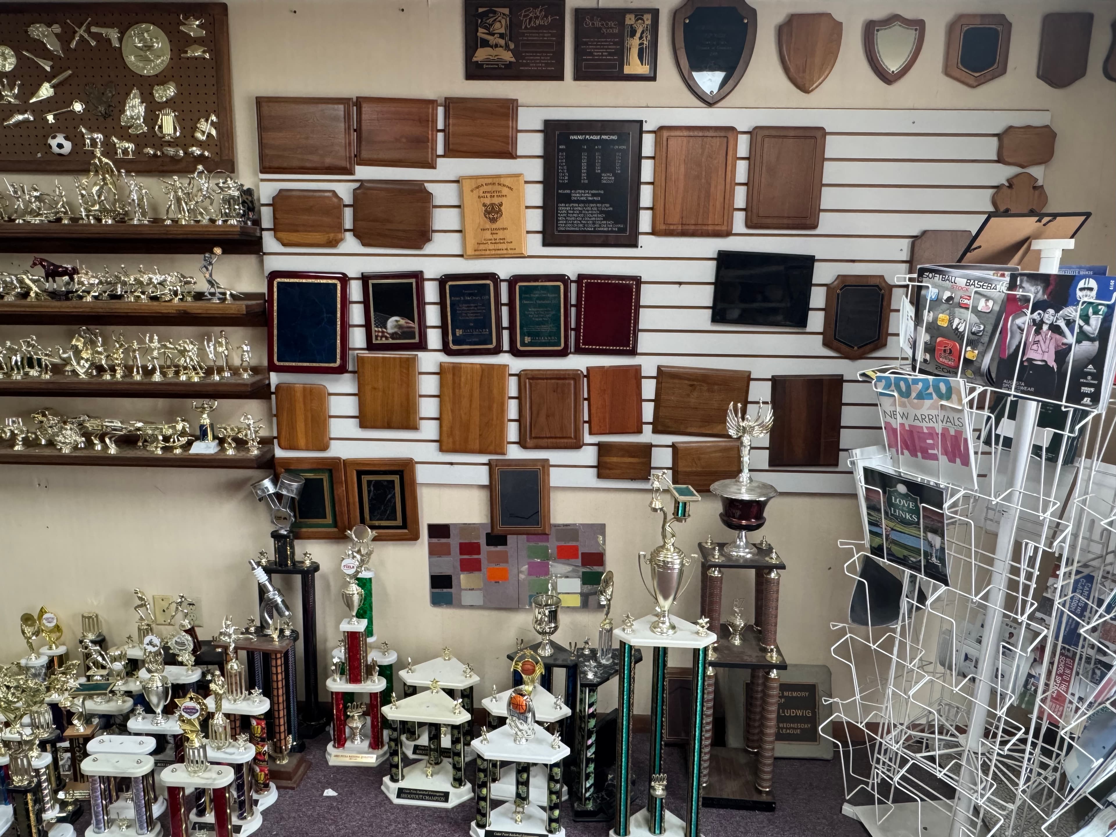 Trophy shop showroom featuring shelves of gold trophies and a wall of wooden plaques.